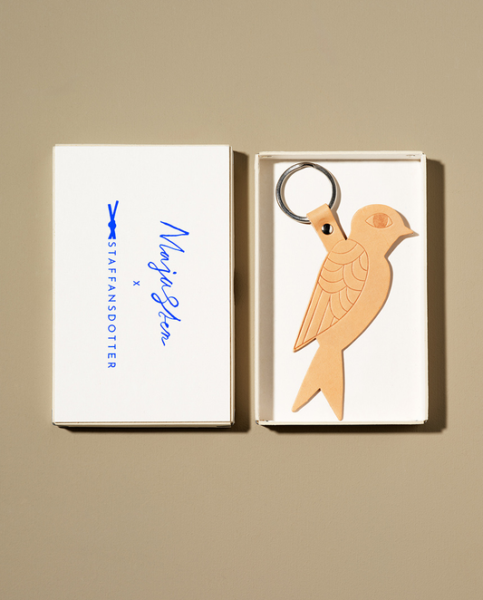 A leather key ring shaped like a bird, displayed in an open cardboard box with an embossed sky-blue logo.