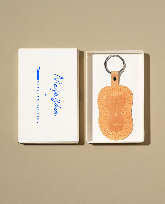 A leather key ring shaped like a monkey's face, presented in a white cardboard box with a sky-blue logo. 